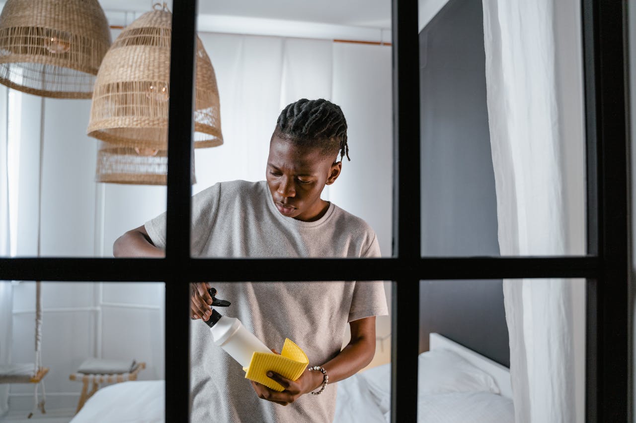 A man with dreadlocks cleans a bedroom using a spray bottle and yellow sponge.